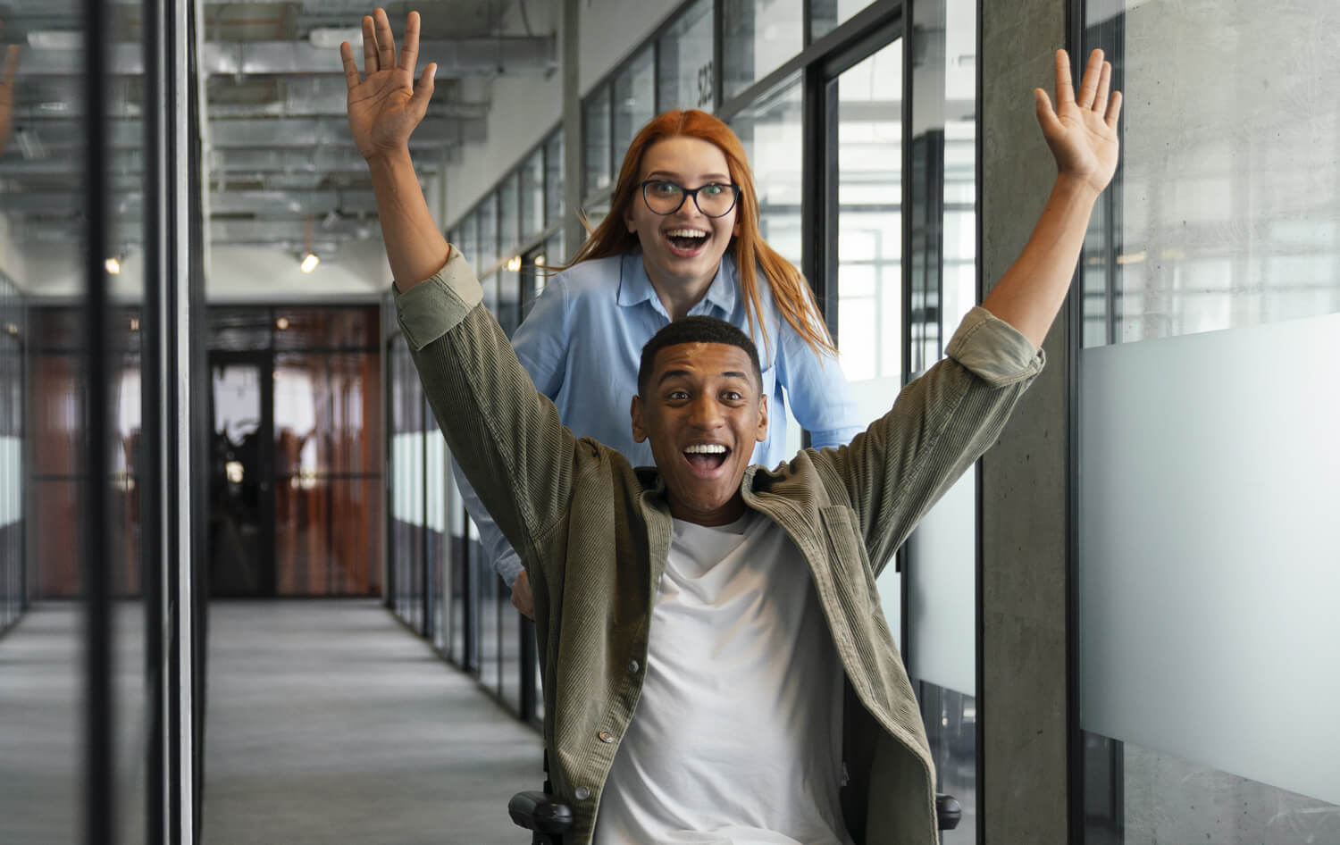 An exuberant young man in a wheelchair with arms raised in  excitement, with a carer woman pushing the wheelchair