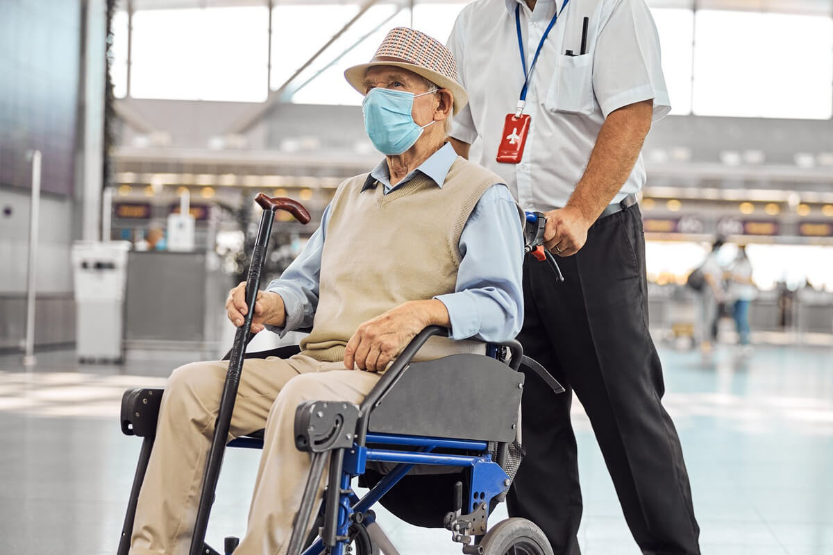 An elderly man wearing a face mask sitting in a wheelchair, pushed by a uniformed attendant at an airport